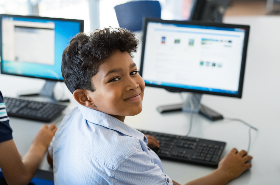 A young elementary school student with dark, curly hair, wearing a light blue striped shirt, sits at a computer in a classroom, looking over his shoulder and smiling directly at the camera. Another computer monitor is visible behind him.